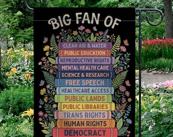 May include: Black garden flag with the text "BIG FAN OF" above a stack of colorful books. Each book lists a different cause, including "CLEAN AIR & WATER" and "DEMOCRACY". The flag is decorated with flowers and butterflies.