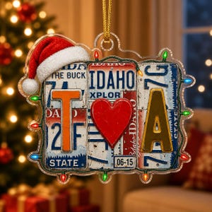 Può includere: Un ornamento decorativo da appendere con tre targhe dell'Idaho affiancate. La lettera T è a sinistra, un cuore rosso al centro e la lettera A a destra. Un cappello da Babbo Natale rosso si trova in alto a sinistra e una serie di luci natalizie colorate incornicia l'intero pezzo.