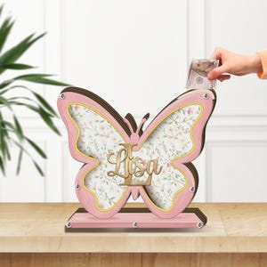Può includere: Un salvadanaio in legno a forma di farfalla con cornice rosa e pannello frontale trasparente. L'interno presenta un motivo floreale e il nome Lisa in lettere dorate. Una fessura sulla parte superiore permette l'inserimento di banconote.