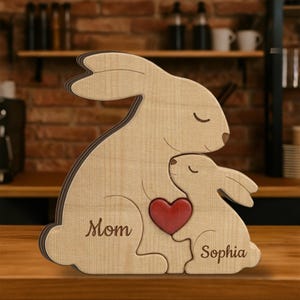 May include: Wooden bunny family sculpture with a red heart. The larger bunny has the word "Mom" engraved, and the smaller bunny has the name "Sophia". The sculpture is made of light-colored wood with a dark brown outline.