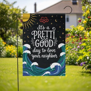 May include: A black garden flag with the text "It's a PRETTI GOOD day to love your neighbour." The flag features a sun, stars, crescent moons, and stylized waves in shades of blue and white. A red heart-shaped knot is at the top.