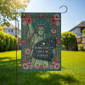 May include: A garden flag featuring the Statue of Liberty holding a stone tablet with the text "democracy dies in silence." The flag has a teal background with floral accents in red and pink. A black metal garden flag holder is attached.