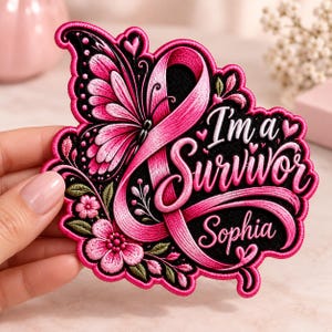 May include: An embroidered patch featuring a pink awareness ribbon, a butterfly with pink and black wings, and floral accents. The text reads I'm a Survivor and the name Sophia in pink script lettering against a black background with small hearts.