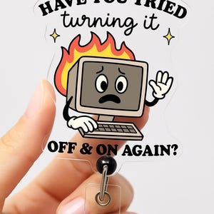 Puede incluir: Un carrete de insignia transparente con un diseño de ordenador de dibujos animados. El ordenador tiene una cara triste y está rodeado de llamas. El texto en la insignia dice "HAVE YOU TRIED turning it OFF & ON AGAIN?"
