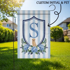 May include: A garden flag with a light blue and white striped pattern. It displays a navy blue shield with the letter "S" and a golden retriever illustration. Blue hydrangea flowers and the text "CUSTOM INITIAL & PET" decorate the bottom of the flag.