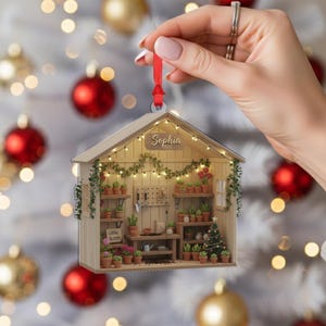 May include: A wooden ornament shaped like a greenhouse, filled with miniature potted plants and a small Christmas tree. The ornament has the name "Sophia" on a sign and is decorated with string lights and the words "Grow Bloom".