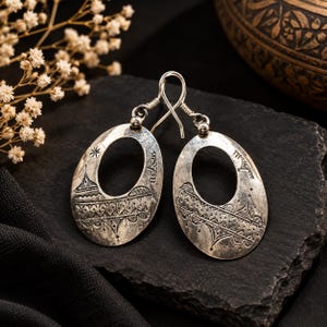 Peut inclure: Une paire de boucles d'oreilles ovales en argent avec une découpe centrale et des motifs gravés complexes. La surface métallique présente une finition oxydée et sombre qui met en valeur les gravures géométriques et linéaires détaillées.