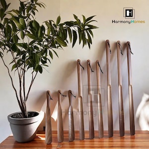 May include: A set of ten wooden shoe horns with leather straps. The shoe horns are various shades of brown and are arranged in a row on a wooden surface. The shoe horns are all different lengths.