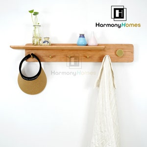 May include: A wooden wall shelf with brass hooks and a round knob. A straw visor and a white towel hang from the shelf. Small vases and a glass bottle with greenery are on the shelf. The logo "Harmony Homes" is visible.