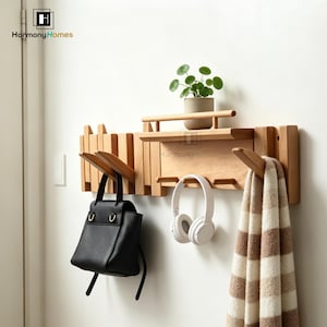 May include: Wooden wall-mounted coat rack with multiple hooks, a shelf, and a small drawer. A black handbag, white headphones, and a striped scarf hang from the hooks. A small potted plant sits on the shelf. The brand name "Harmony Homes" is visible.