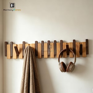 May include: A wooden wall-mounted coat rack with alternating light and dark wood slats. A brown blanket and brown headphones hang from the hooks. The rack is mounted on a white wall.