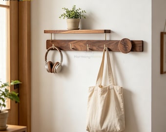 May include: A wooden wall-mounted shelf with a lower bar featuring brass hooks. A small potted plant sits on the shelf above. A pair of headphones and a canvas tote bag hang from the hooks. The wall is white, and the wood is a rich brown.
