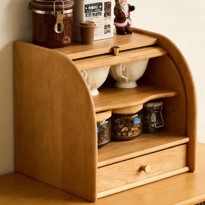 May include: A light-colored wooden bread box with a roll-top design. It features multiple shelves, a drawer, and glass jars filled with coffee beans. The top shelf holds a brown jar, a small cup, and a decorative Santa Claus figurine.