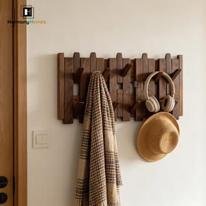 May include: A dark brown wooden coat rack with multiple hooks, holding a plaid scarf, a pair of headphones, and a straw hat. The rack is mounted on a white wall. The logo "Harmony Homes" is in the top left corner.
