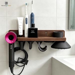 Könnte beinhalten: Ein wandmontiertes Badezimmerregal aus dunklem Holz mit einer Halterung für einen Haartrockner mit pinker Düse, zwei Steckplätzen für elektrische Zahnbürsten, einer Ablage für Pflegeprodukte, einem schwarzen Haken und einem schwarzen Becher.