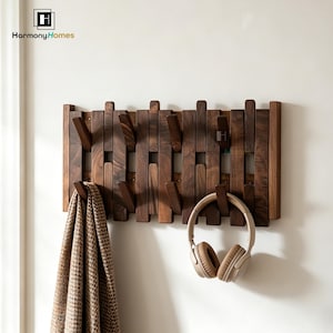 May include: A wooden wall-mounted coat rack with multiple hooks. The rack is made of dark brown wood and has a unique design with vertical and horizontal wooden slats. A brown and tan patterned scarf and a pair of tan headphones hang from the hooks.