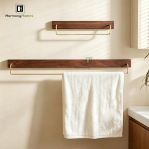 May include: Two wooden towel racks with brass bars are mounted on a light-colored wall. The top rack is smaller, while the bottom rack holds a folded white towel. The brand name "Harmony Homes" is visible in the top left corner.