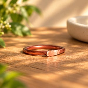 May include: A circular ring crafted from polished copper, featuring a smooth, rounded band that transitions into a single, flattened, and hammered end section. The metal exhibits a warm, reddish-brown metallic luster with visible texture.