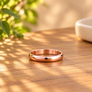 Puede incluir: Un anillo de oro rosa pulido con una banda lisa y redondeada. La superficie metálica es altamente reflectante, mostrando un brillo metálico cálido. El anillo presenta un diseño circular simple y continuo sin piedras, grabados ni patrones.
