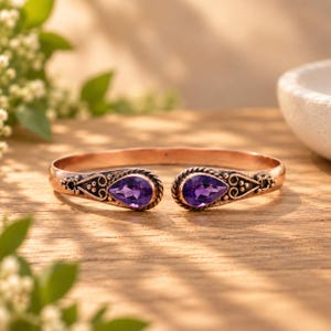 Può includere: Un bracciale a fascia in rame dal design aperto, caratterizzato da due pietre preziose di ametista viola a forma di goccia alle estremità. La fascia metallica presenta complessi motivi a volute e perline attorno a ogni pietra.