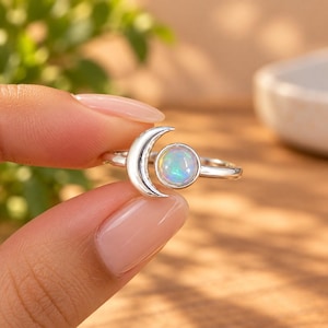 May include: A silver ring featuring a crescent moon shape next to a round, iridescent opal cabochon. The band is thin and polished, showcasing the reflective quality of the stone which displays flashes of blue, green, and orange light.