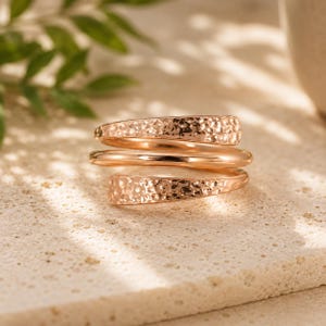 May include: A rose gold bypass ring featuring a multi-layered design with two textured, hammered bands flanking a smooth, polished central band. The metal has a warm, metallic sheen and a continuous wrap-around structure that creates a layered appearance.
