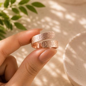 May include: A rose gold-toned open ring featuring a textured, hammered surface finish. The band is constructed with two overlapping ends that do not meet, creating a bypass style design with a consistent metallic sheen across the entire exterior.
