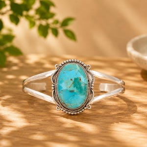 May include: A silver cuff bracelet featuring a large, oval-shaped turquoise stone at the centre. The stone displays natural blue and green hues with brown matrix veining. The setting is framed by a decorative beaded and twisted metal border.