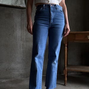 May include: Medium-wash blue denim jeans with a high waist and straight leg, featuring a frayed hem. The jeans are worn with a white, cropped top and black patent leather flats. The background is a neutral, industrial setting.