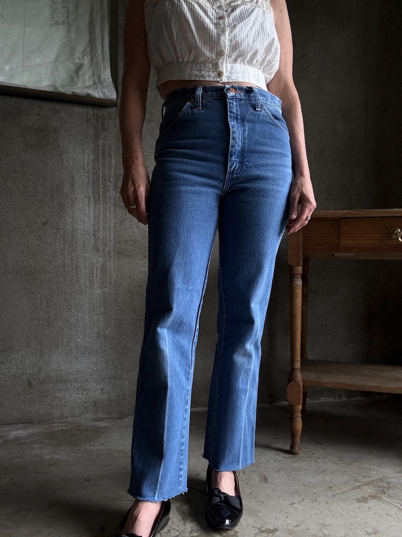 May include: Medium-wash blue denim jeans with a high waist and straight leg, featuring a frayed hem. The jeans are worn with a white, cropped top and black patent leather flats. The background is a neutral, industrial setting.