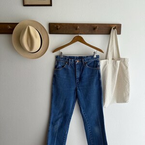 May include: Blue denim jeans hanging on a wooden hanger. A straw hat and a canvas tote bag with a dotted pattern are also visible. A framed picture hangs above the hat.