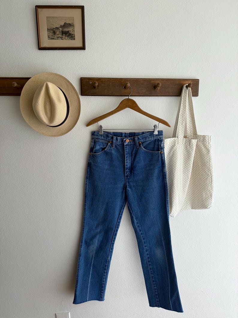 May include: Blue denim jeans hanging on a wooden hanger. A straw hat and a canvas tote bag with a dotted pattern are also visible. A framed picture hangs above the hat.