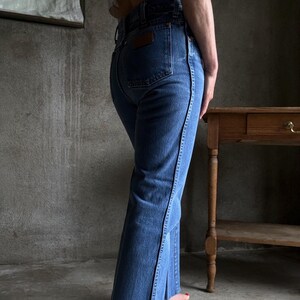 May include: Blue denim jeans with a flared leg and a dark brown leather patch on the back pocket. The jeans have a classic five-pocket design and a black seam detail along the side of the leg.