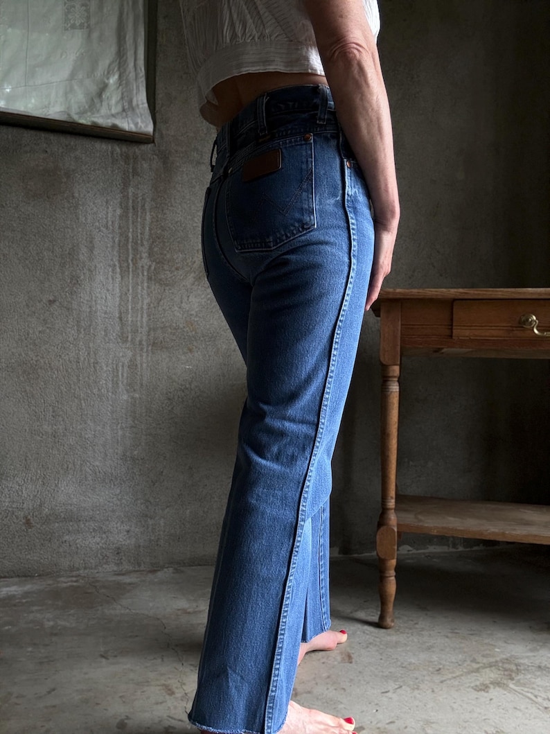 May include: Blue denim jeans with a flared leg and a dark brown leather patch on the back pocket. The jeans have a classic five-pocket design and a black seam detail along the side of the leg.