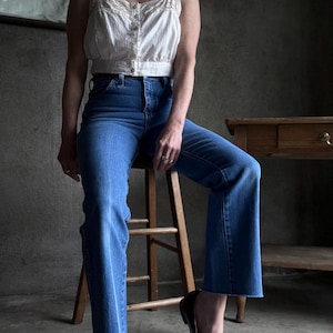 May include: A white, vertically striped crop top with lace trim and button details paired with blue jeans. The jeans have a flared leg and are cuffed at the ankle. Black flats with bows complete the outfit.