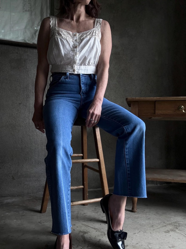 May include: A white, vertically striped crop top with lace trim and button details paired with blue jeans. The jeans have a flared leg and are cuffed at the ankle. Black flats with bows complete the outfit.