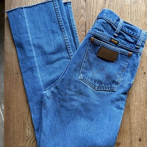 May include: Light blue denim jeans with a Wrangler brand patch on the back pocket. The jeans have a classic five-pocket design and a straight leg cut. The jeans are made of a durable denim material.