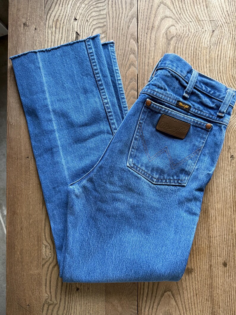 May include: Light blue denim jeans with a Wrangler brand patch on the back pocket. The jeans have a classic five-pocket design and a straight leg cut. The jeans are made of a durable denim material.