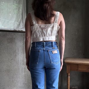 May include: A person wearing a straw hat with a black band, a cream-colored lace-trimmed crop top, and blue denim jeans. The jeans have a Wrangler patch on the back pocket. The outfit is set against a grey background.