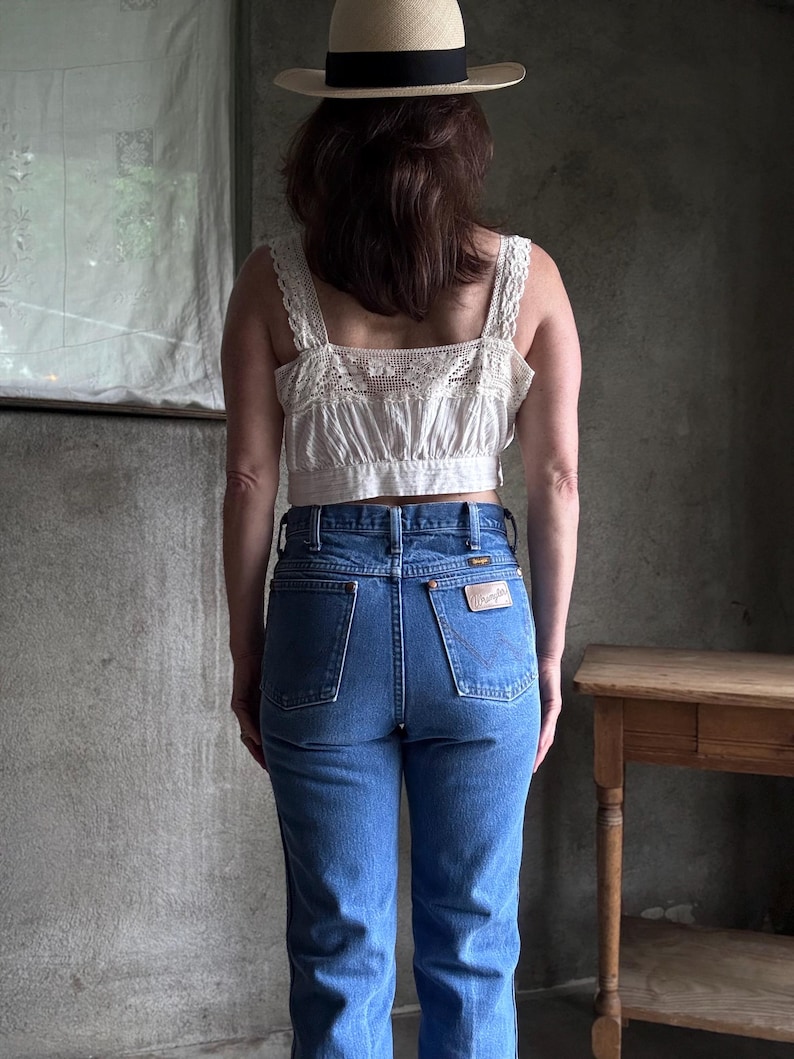 May include: A person wearing a straw hat with a black band, a cream-colored lace-trimmed crop top, and blue denim jeans. The jeans have a Wrangler patch on the back pocket. The outfit is set against a grey background.