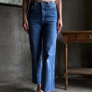 May include: Blue denim jeans with a high waist and flared leg. The jeans have a classic five-pocket design and a button and zip closure. The jeans are a medium wash and appear to be vintage.