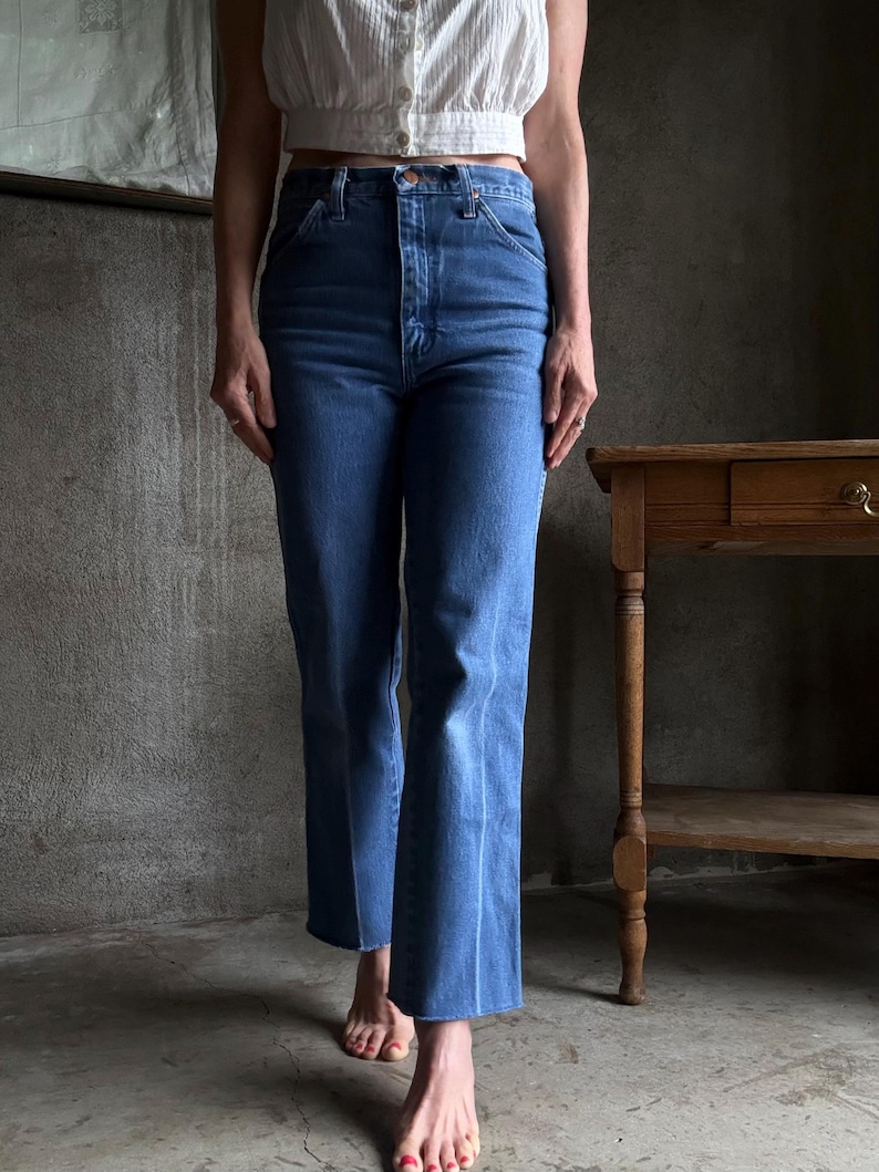 May include: Blue denim jeans with a high waist and flared leg. The jeans have a classic five-pocket design and a button and zip closure. The jeans are a medium wash and appear to be vintage.