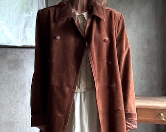 May include: A brown suede coat with a double-breasted design. The coat has a collared neckline and long sleeves. The model is wearing a white hat and a white dress underneath the coat. The coat has two rows of buttons.