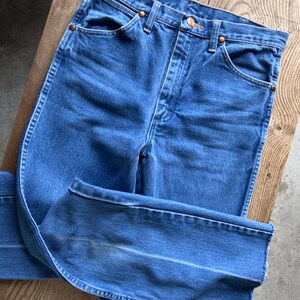 May include: Blue denim jeans with a classic five-pocket design. The jeans have a high waist and a straight leg cut. Copper rivets and button detail. The jeans are folded on a wooden surface.