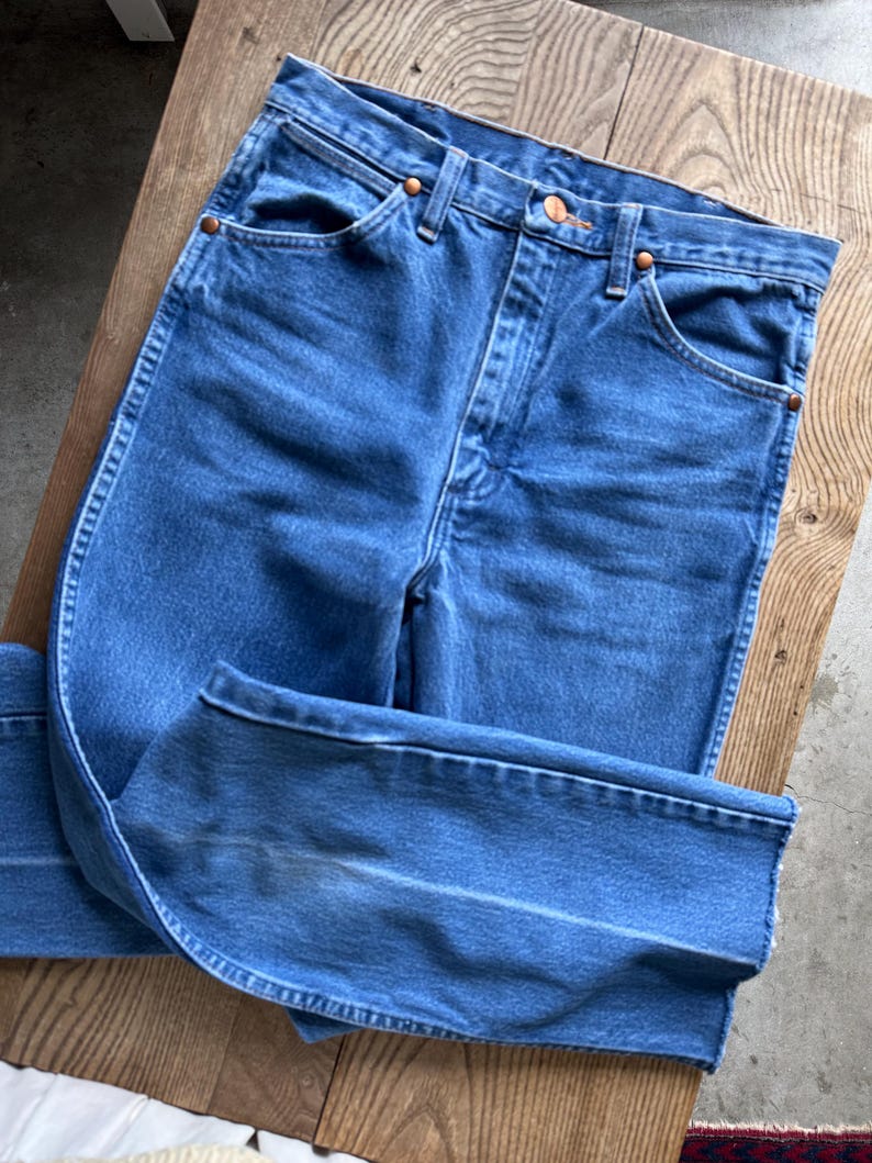 May include: Blue denim jeans with a classic five-pocket design. The jeans have a high waist and a straight leg cut. Copper rivets and button detail. The jeans are folded on a wooden surface.