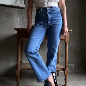 May include: Light-wash blue jeans with a high waist and straight leg, featuring a raw hem. The jeans are worn with a white, button-up crop top and black patent leather loafers with bows. A small wooden table is in the background.