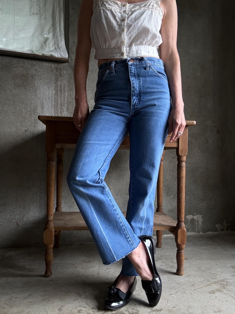 May include: Light-wash blue jeans with a high waist and straight leg, featuring a raw hem. The jeans are worn with a white, button-up crop top and black patent leather loafers with bows. A small wooden table is in the background.