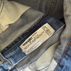 May include: Close-up of blue denim Wrangler jeans. The jeans have a white label with the brand name and washing instructions. The jeans are size 30 x 36. The jeans are made of 100% cotton.