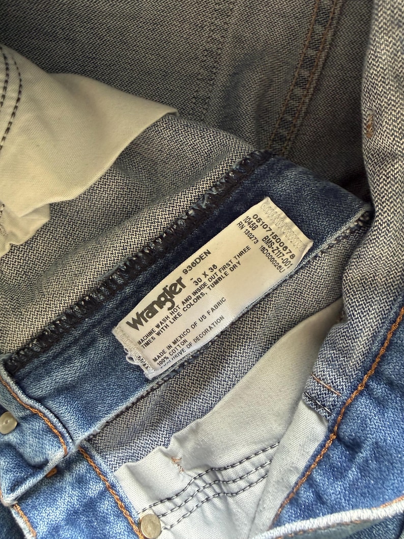 May include: Close-up of blue denim Wrangler jeans. The jeans have a white label with the brand name and washing instructions. The jeans are size 30 x 36. The jeans are made of 100% cotton.