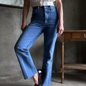 May include: Blue denim jeans with a high waist and flared leg. The jeans have a classic five-pocket design and a light wash. The model is wearing a white top and black shoes. The jeans are a vintage style.